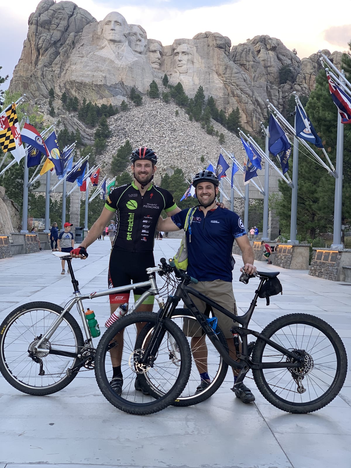 Two riders with their bikes at Mount Rushmore, state flags lining the avenue behind them.
