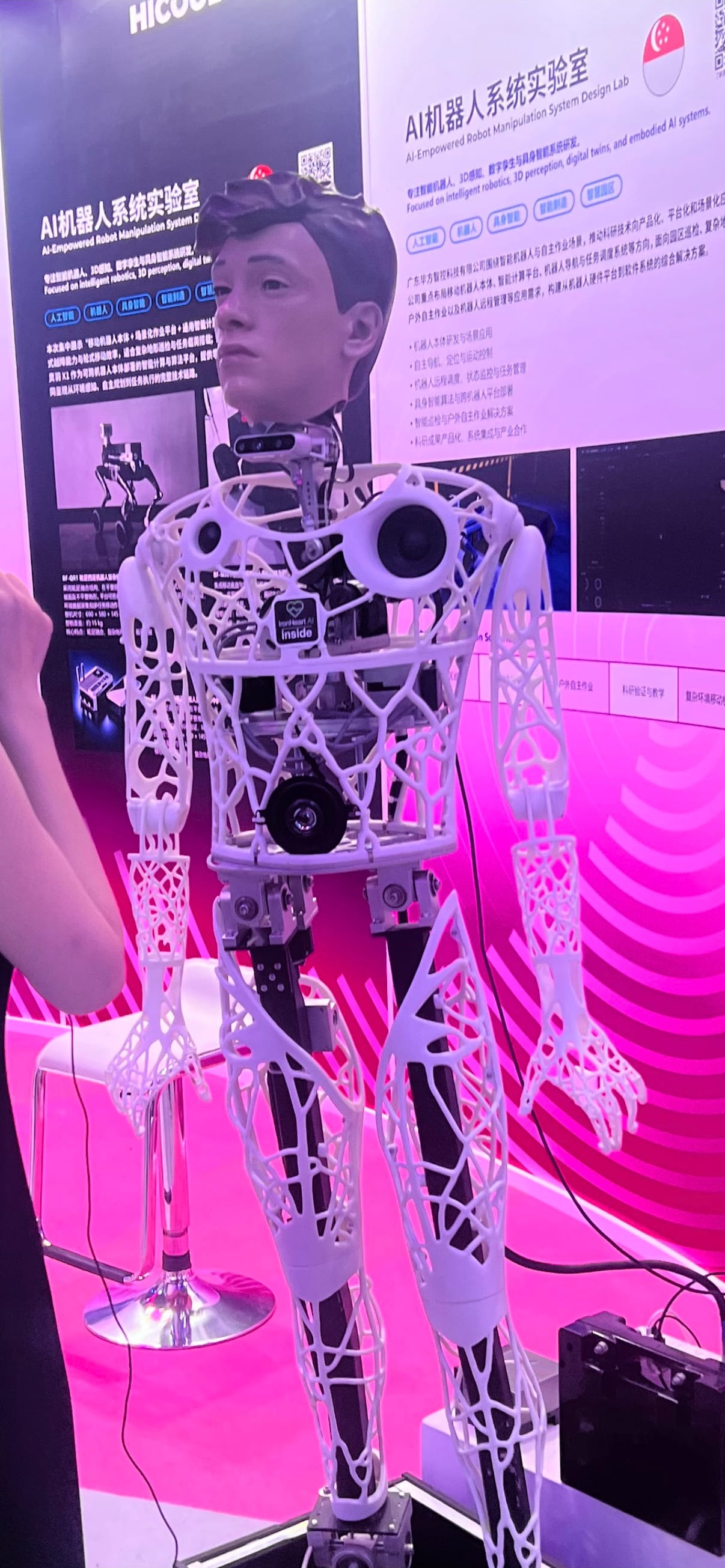 A life-size humanoid robot with a lattice-printed white body and a realistic human face at an exhibit booth.