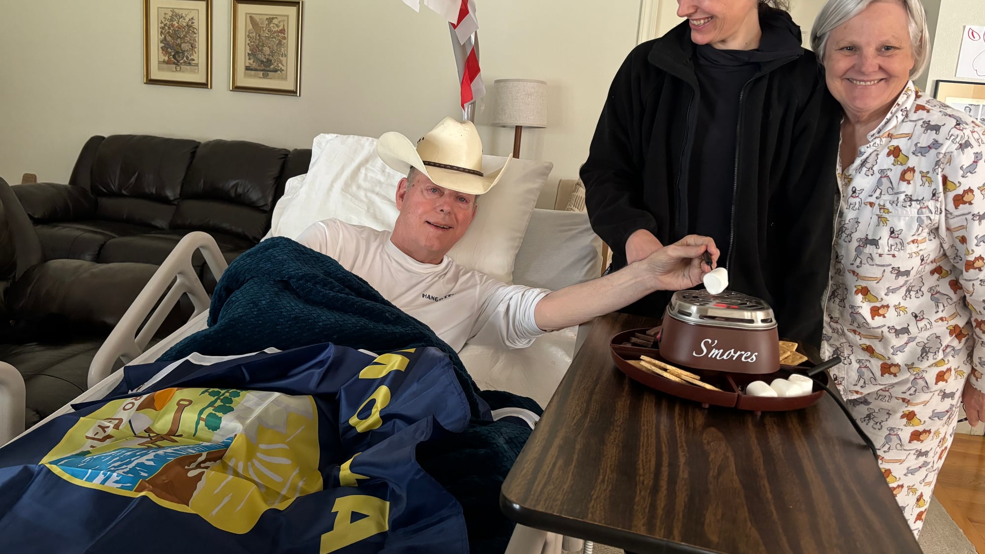 John's dad in a hospital bed at home wearing a cowboy hat, a Montana state flag across the bed, reaching for a marshmallow at a s'mores maker while family stand beside him, smiling.