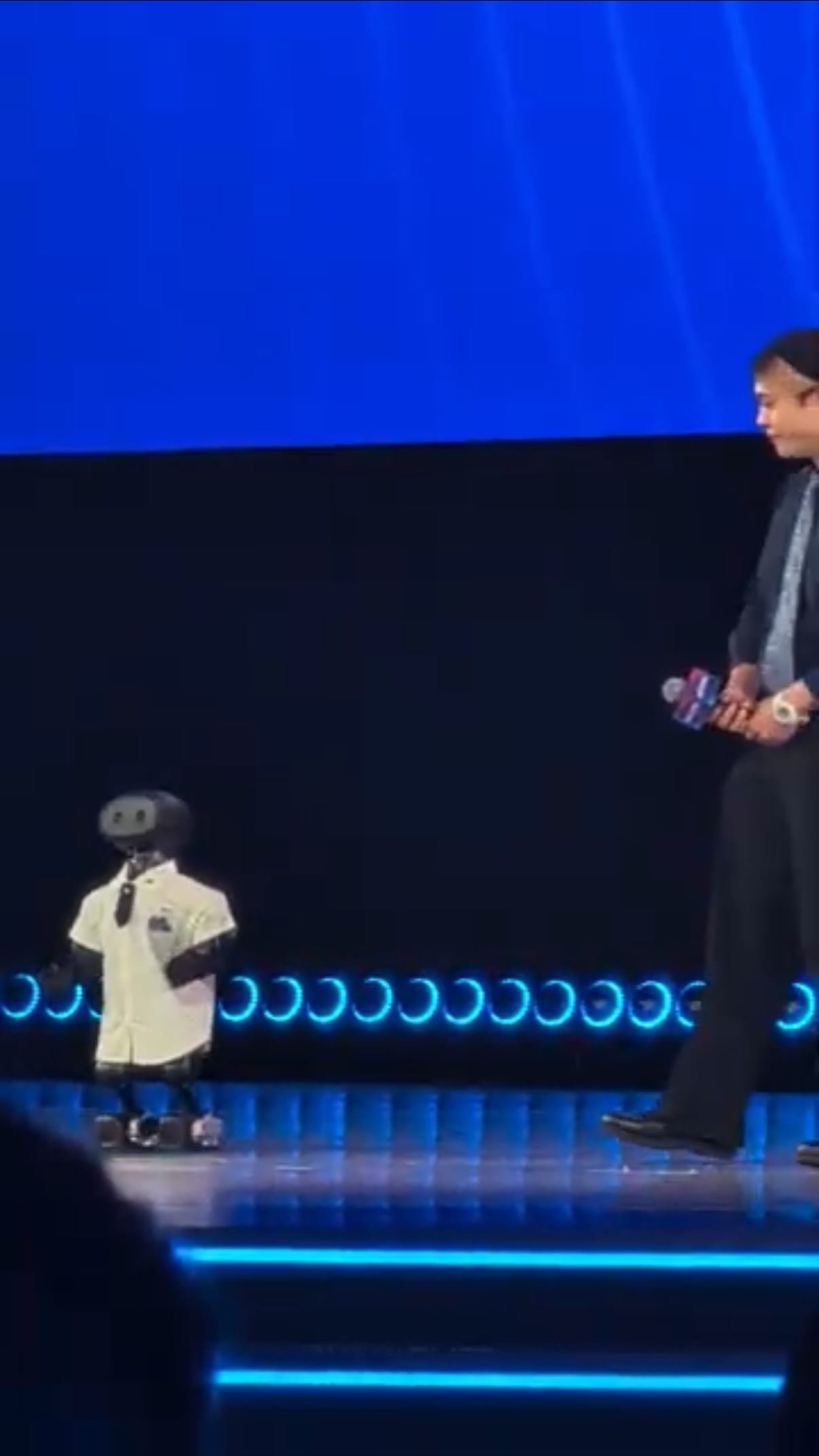 A small humanoid robot in a white shirt standing on a stage beside its founder.